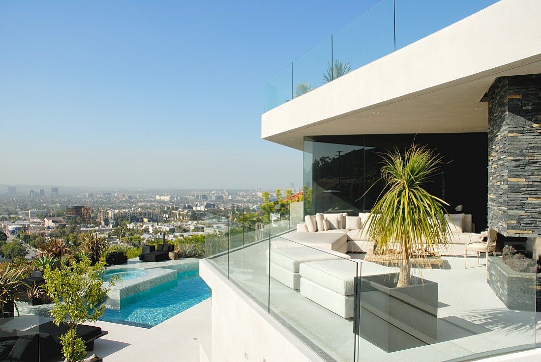 Contemporary house with white outdoor furniture on terrace with glass balustrade; pool and urban panorama in background