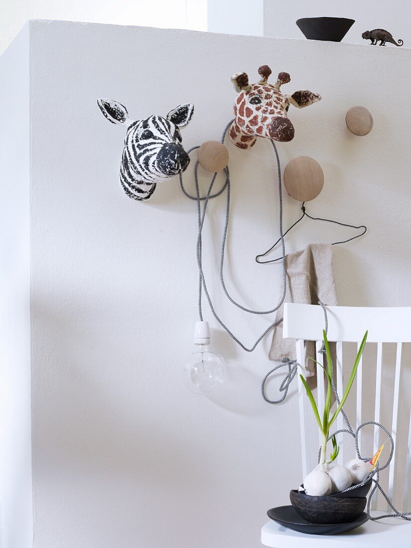 Safari-Tierköpfe aus Pappmache an weißer Wand mit Kleiderhaken und schwarze Schalen auf weißem Holzstuhl