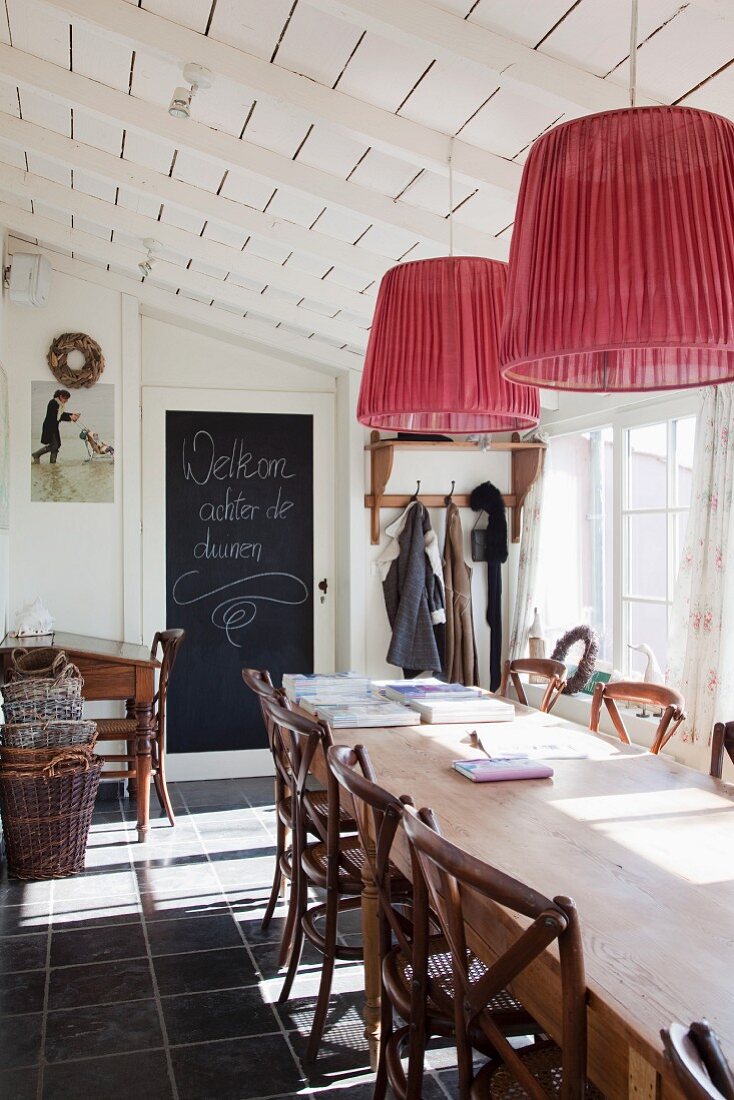 Pendant lamps with red lampshades above rustic wooden chairs in sunny dining area with greeting written on blackboard panel on door