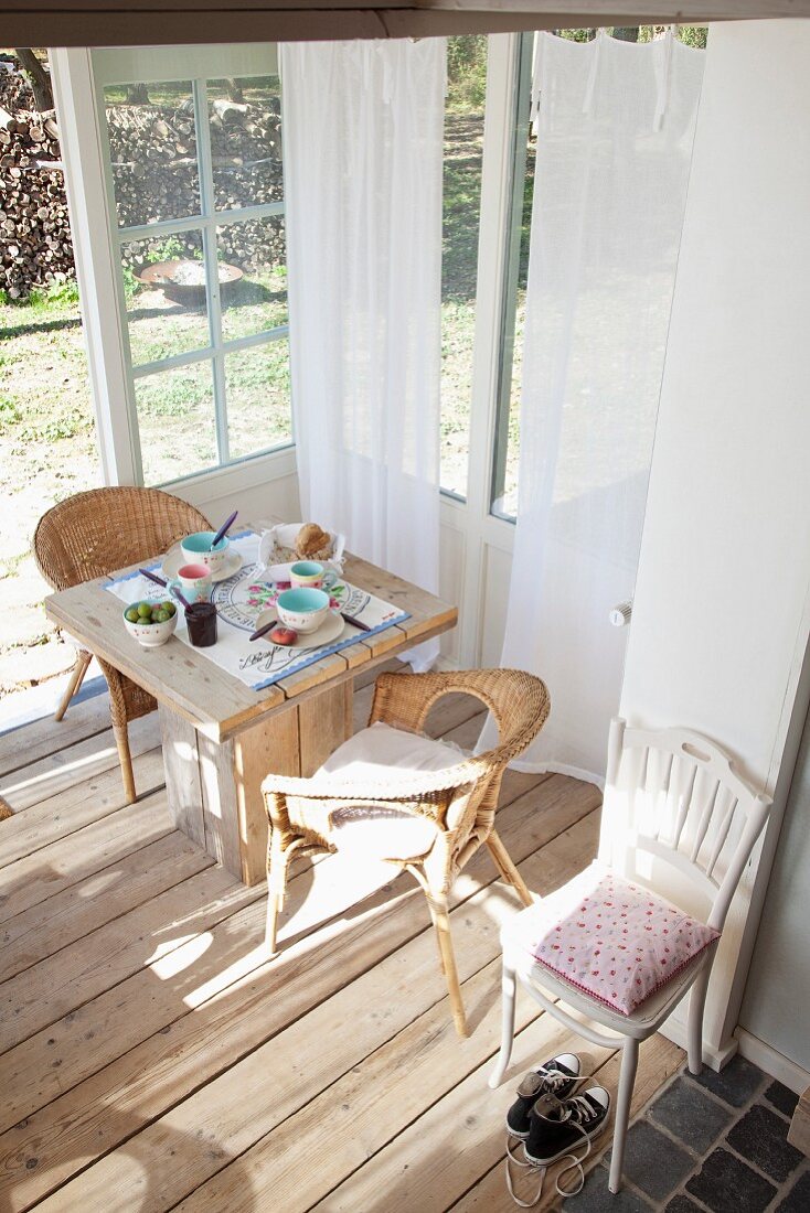 Gedeckter Frühstückstisch mit Korbstühlen in sonnigem Wintergartenanbau mit naturbelassenem Dielenboden