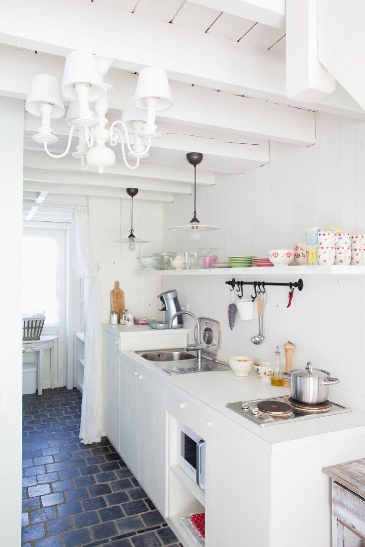 Kitchen counter in white shabby-chic kitchen with antique pendant lamps and chandelier with lampshades hanging from exposed wood-beamed ceiling