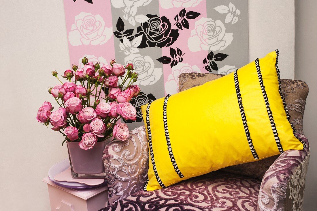 A purple velvet chair with a yellow cushion, vase of pink roses and a stencil background