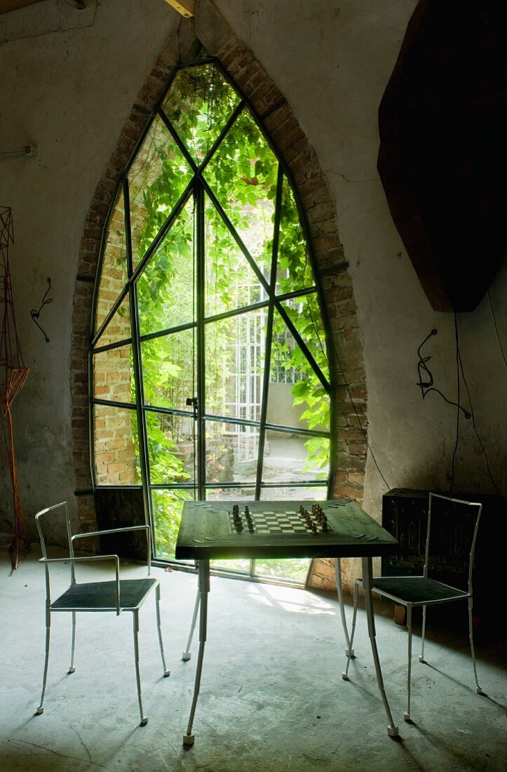 Schachtisch und filigrane Metallstühle vor Spitzbogen mit Sprossentür in künstlerischer Ausführung und Blick in Garten