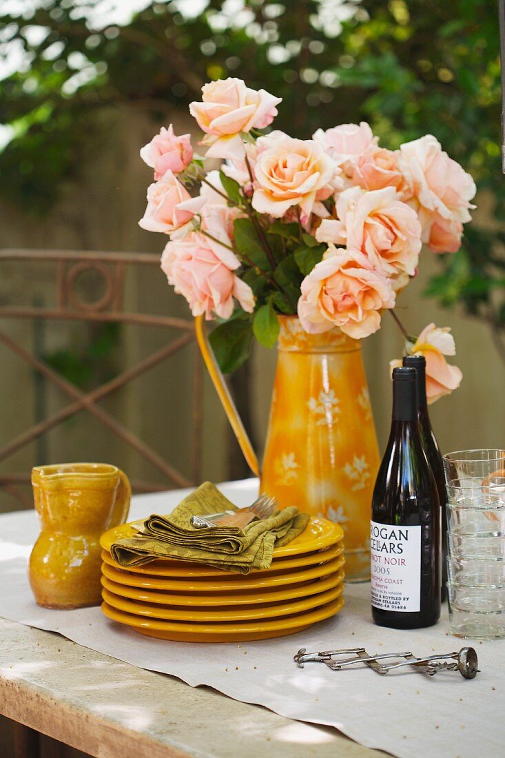 Tellerstapel, lachsfarbene Rosen und Weinflaschen auf einem Tisch im Freien