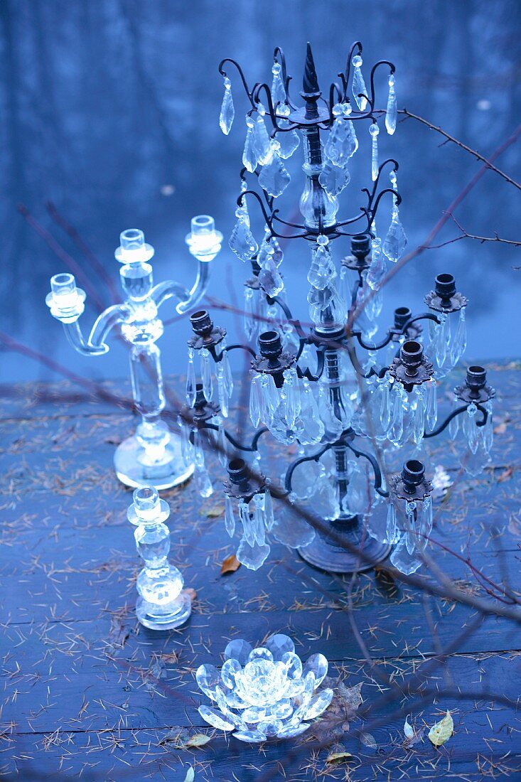 Verschieden grosse Kerzenleuchter aus Glas und Kerzenständer mit Glasschmuck auf Holzdeck am Seeufer