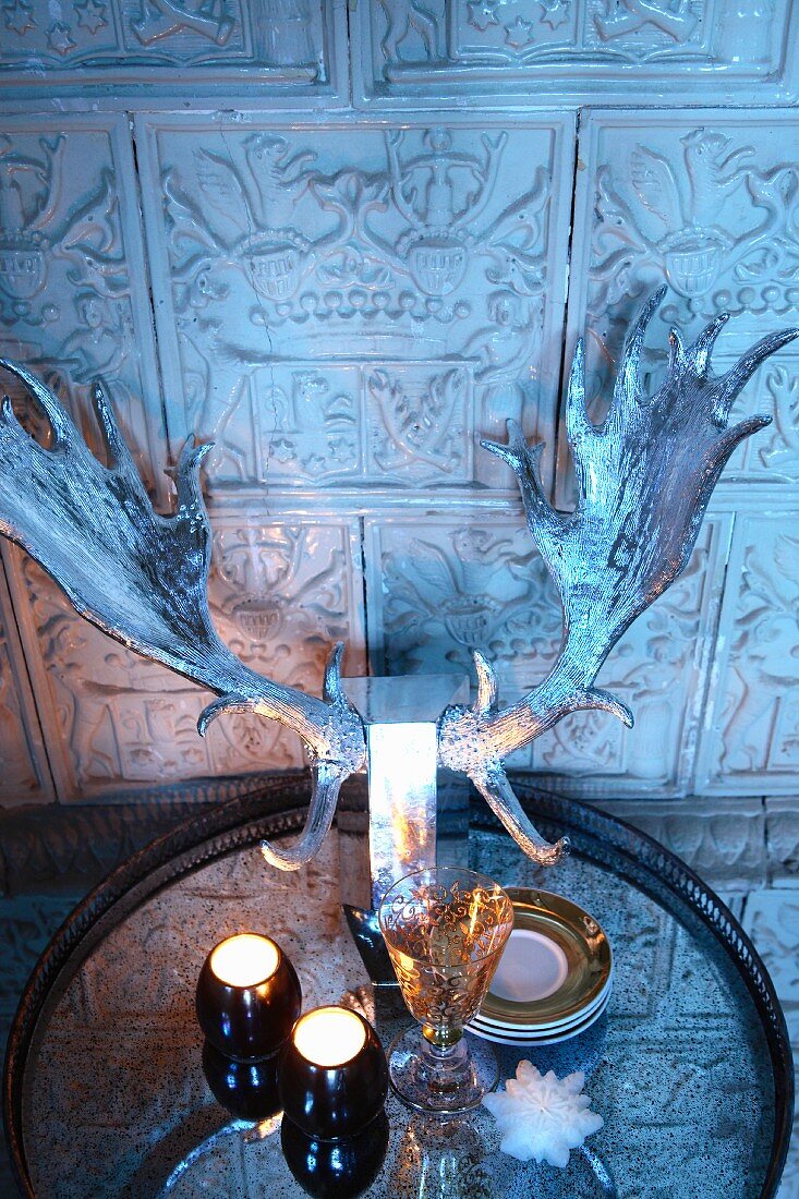 Teelichter und versilbertes Hirschgeweih auf Tablett vor gefliester Wand mit antiken Motiven