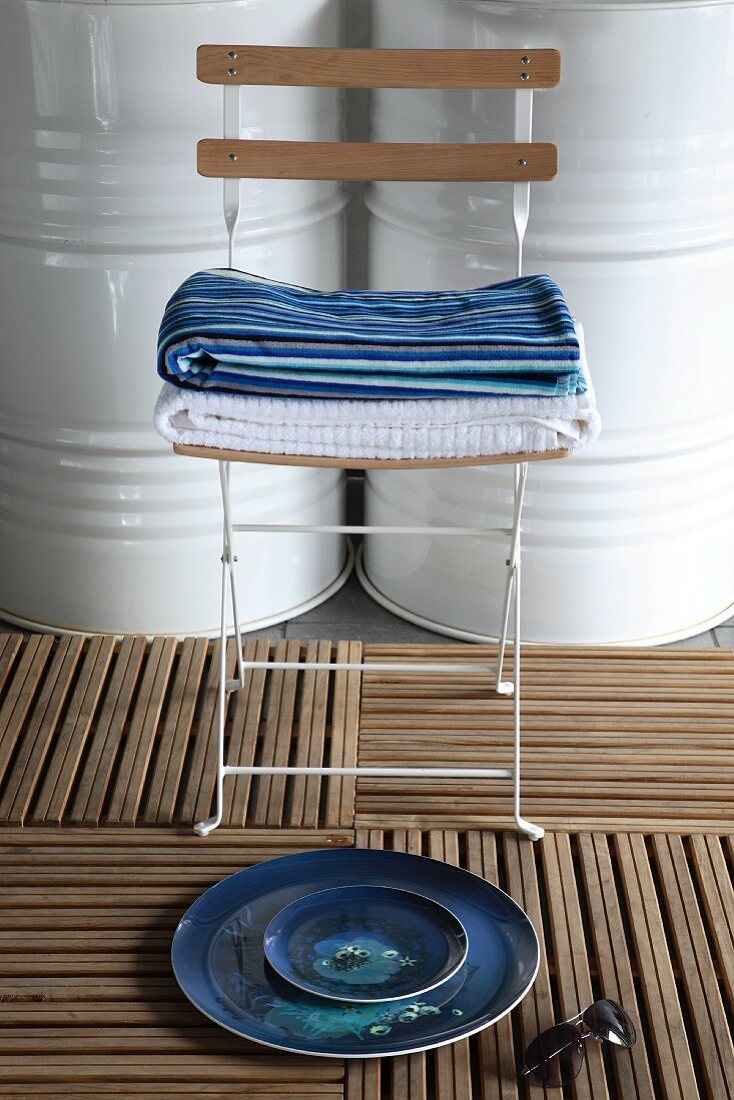 Still-life arrangement of plates on decking tiles in front of stacked towels on folding chair and white-painted metal barrels
