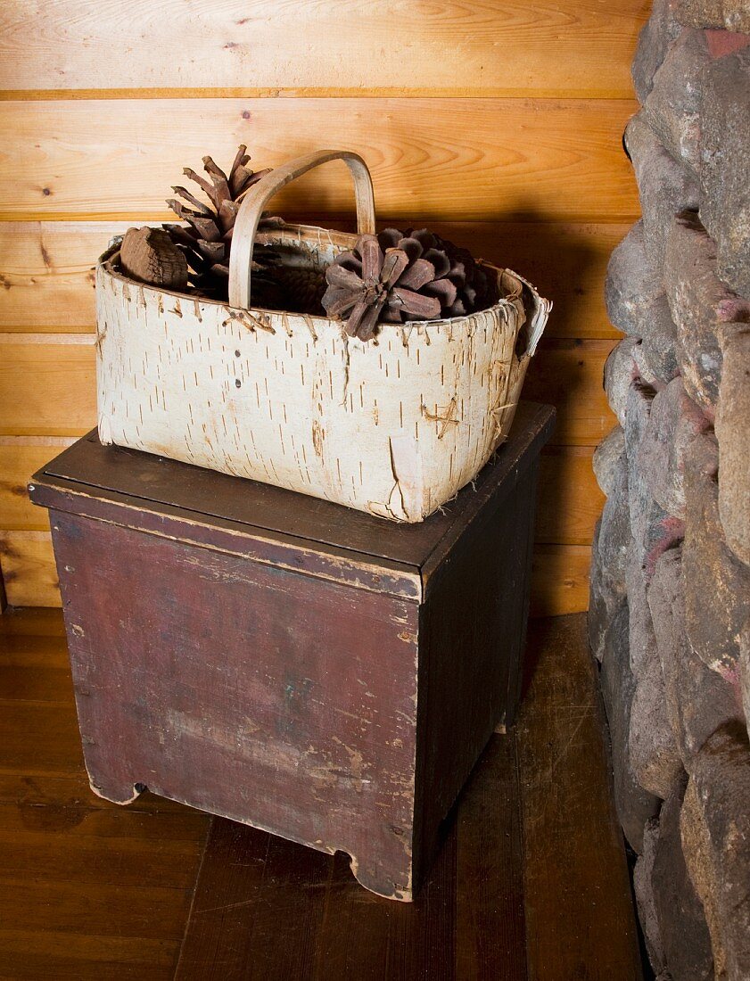 Selbst gemachter Korb aus Birkenrinde mit Pinienzapfen auf schlichtem Holzhocker vor Holzwand in Zimmerecke