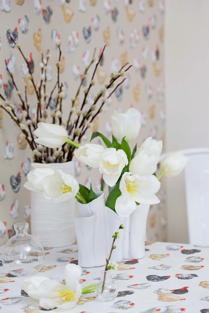 Vases of white tulips and pussy willow on tablecloth with Easter pattern of hens