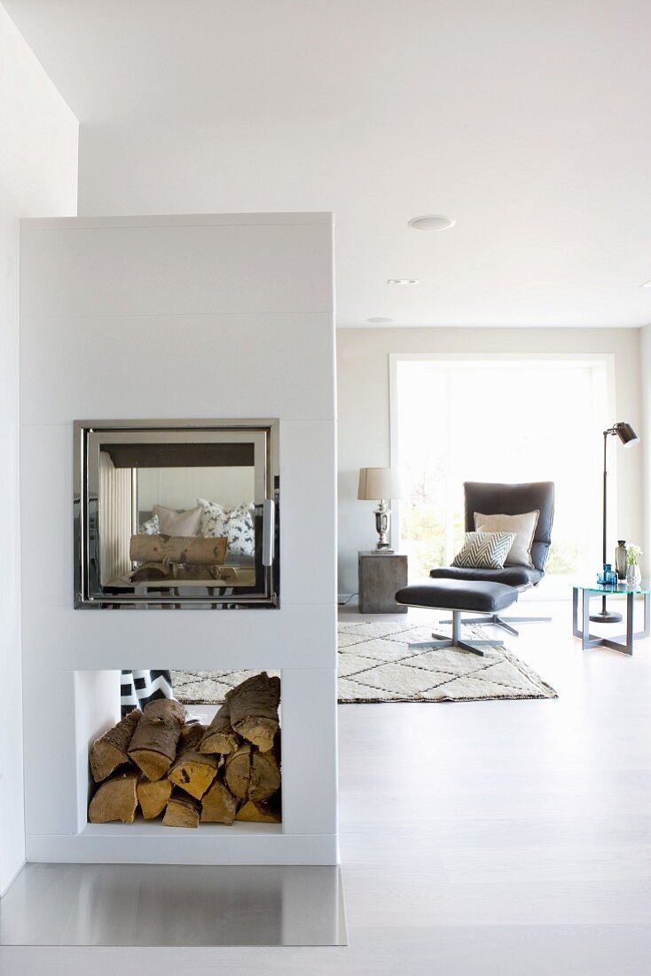 Half-height, masonry fireplace with double-sided firebox and firewood store; easy chair in seating area beyond