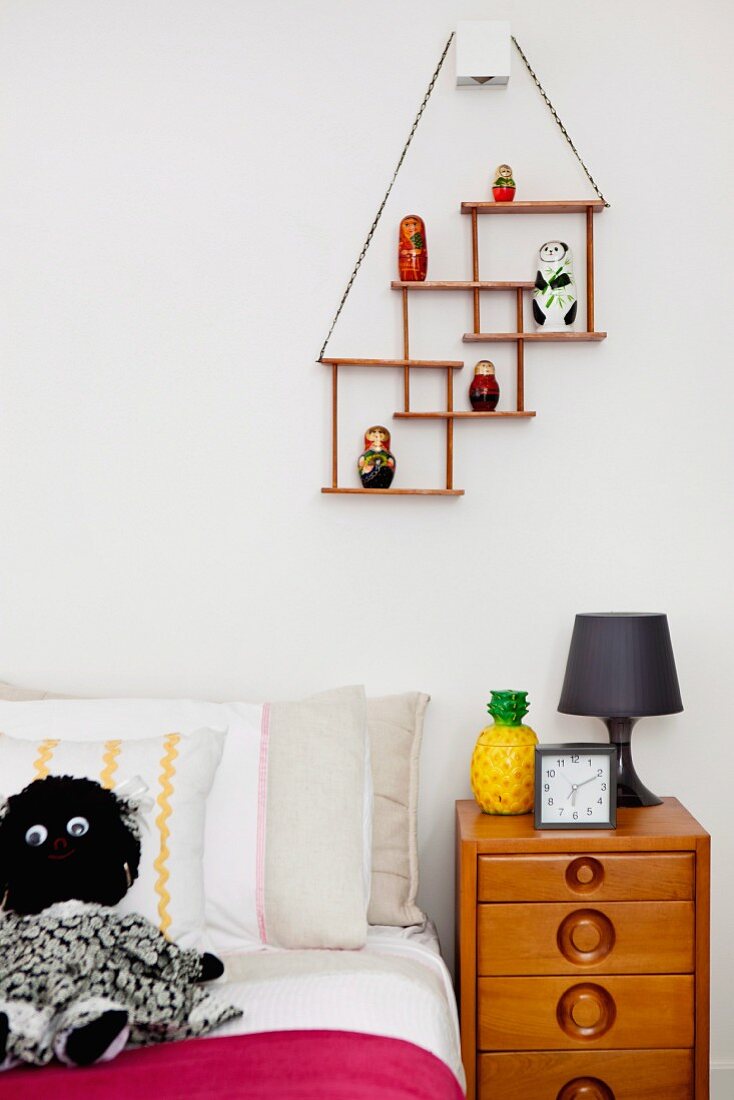 Jugendbett mit schwarzweisser Kuschelpuppe und Retro Holzkommode; an der Wand eine Art minimalistischer Setzkasten mit Figürchen