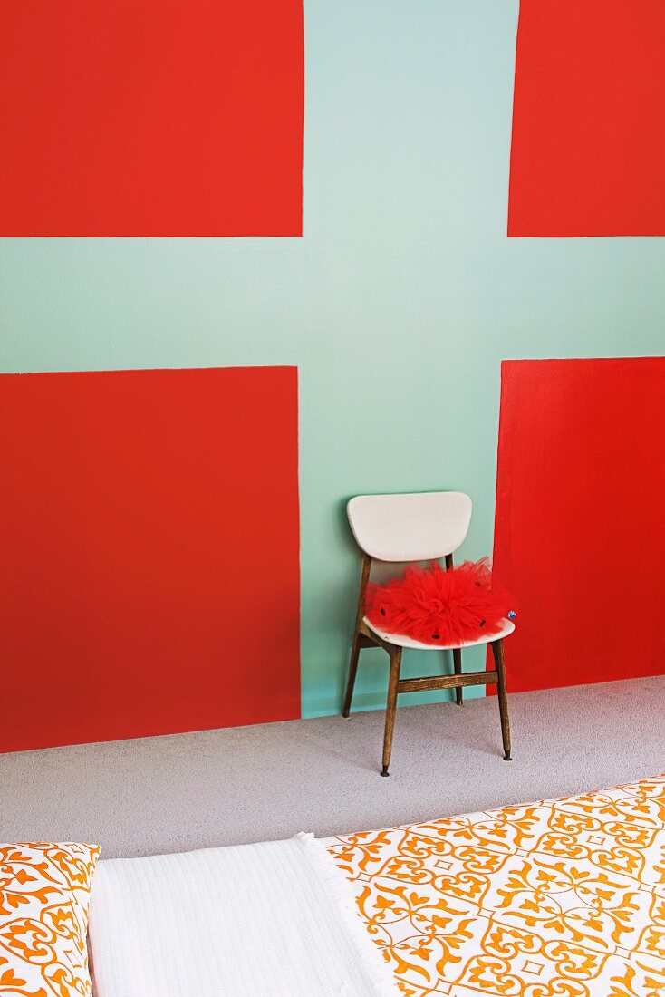 Stuhl mit roter Dekoware an bemalter Wand mit grafischem Muster in Rot und Türkis, davor Bett mit Ornamentmuster auf Bettwäsche