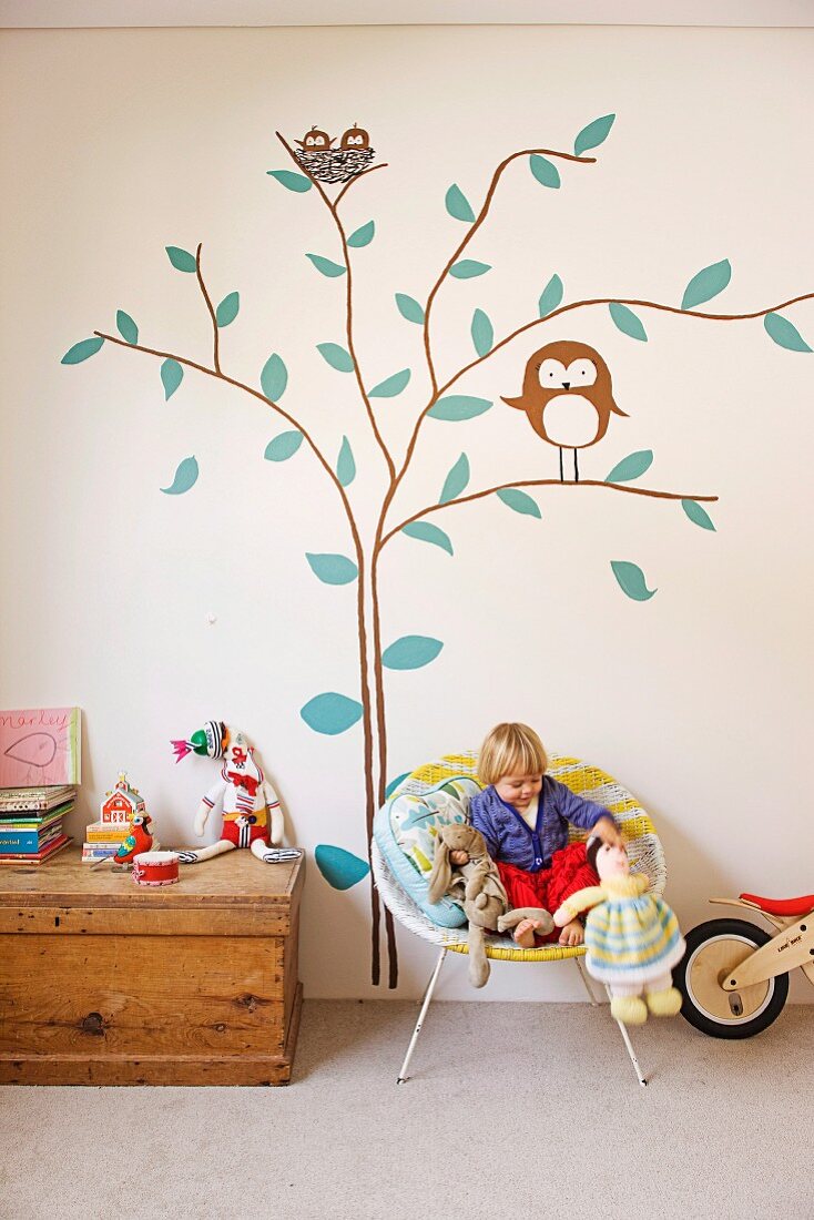 Kind mit Stofftieren auf Schalenstuhl aus Geflecht und rustikale Truhe vor Wand mit gemalten Baum