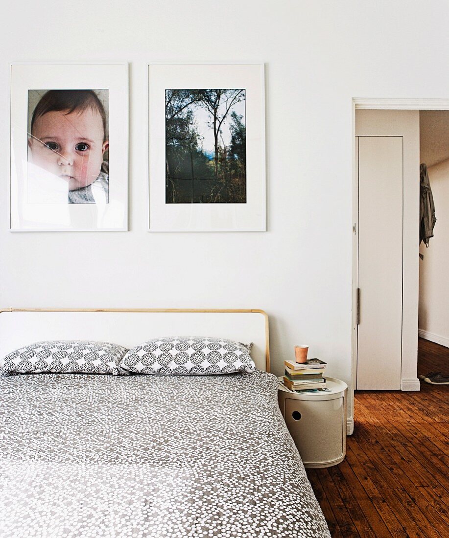 Weissgrau gemusterte Tagesdecke und Kissen auf Doppelbett, vor Wand mit Fotos, seitlich offene Tür und Blick in Gang