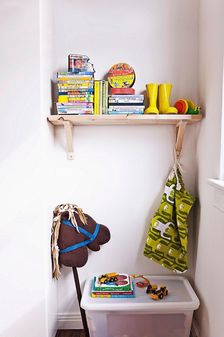 Books and yellow Wellingtons on wooden shelf in niche with hobby horse and toy box on floor