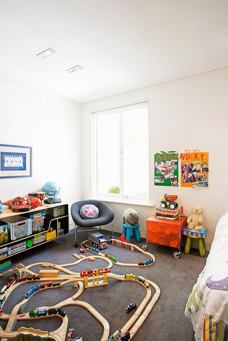 Modell Eisenbahn aus Holz auf Boden, an der Wand offenes Sideboard mit Spielsachen und grau bezogener Sessel in Zimmerecke am Fenster