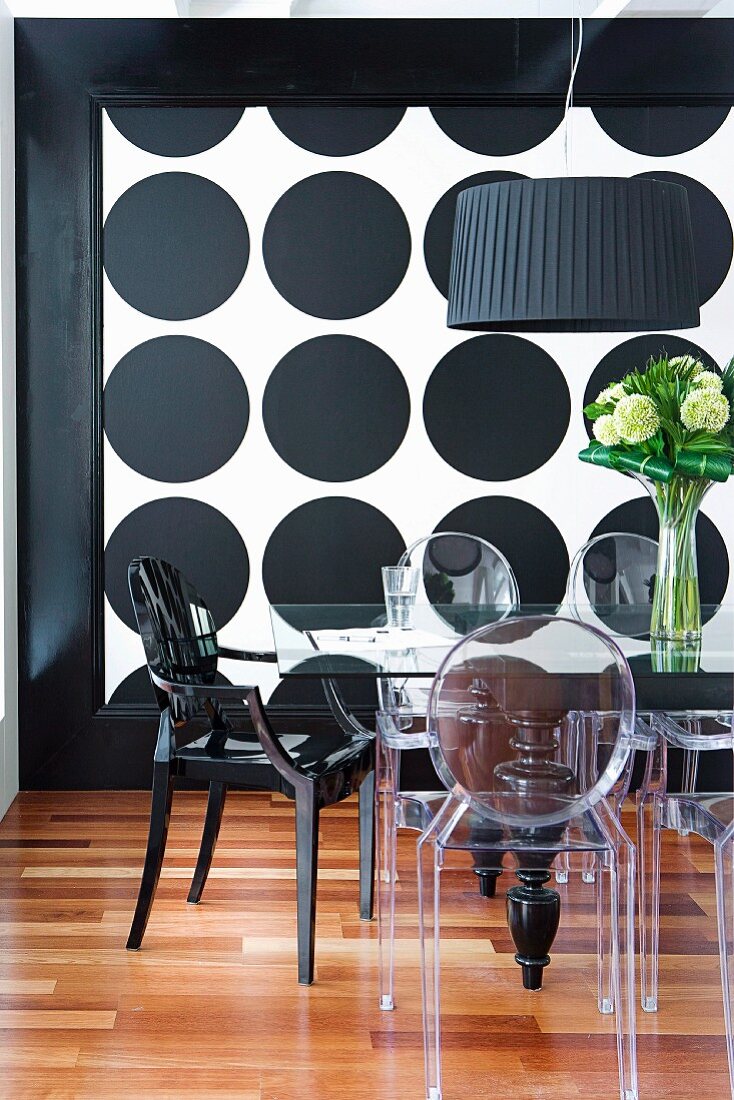 Transparent and black, plastic designer chairs around glass table in front of wall with pattern of large black circles on white background