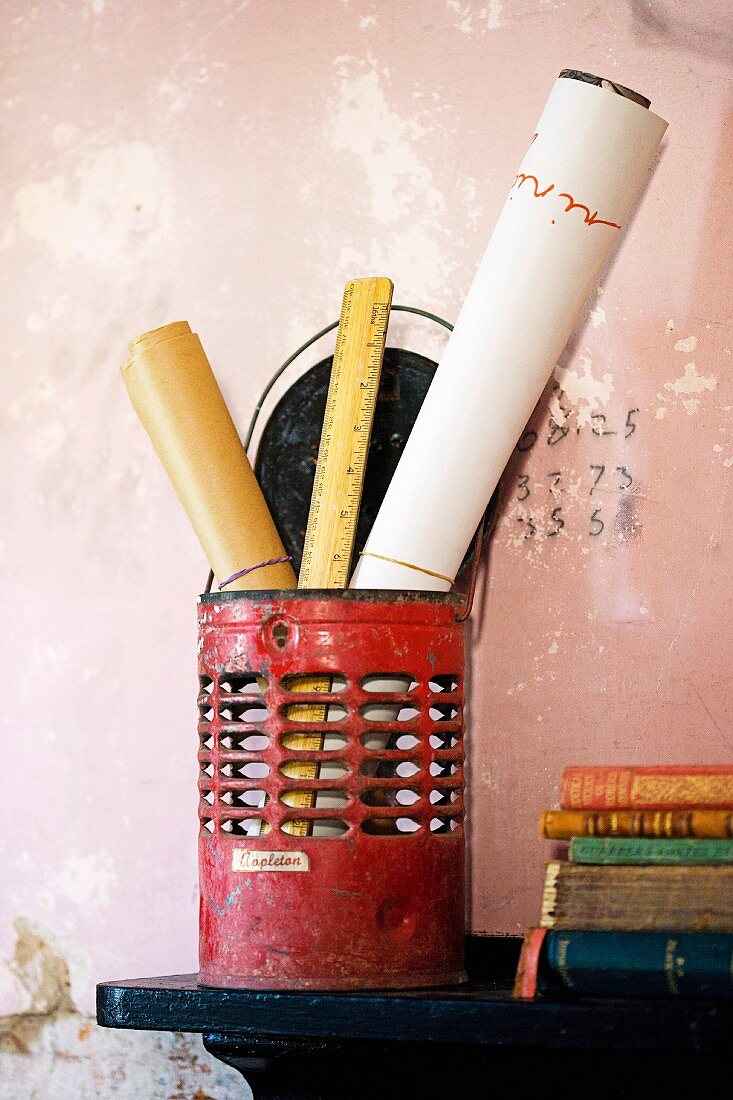 Rolls of paper and rulers in old, red metal container next to stack of books on bracket mounted on wall with peeling pink paint