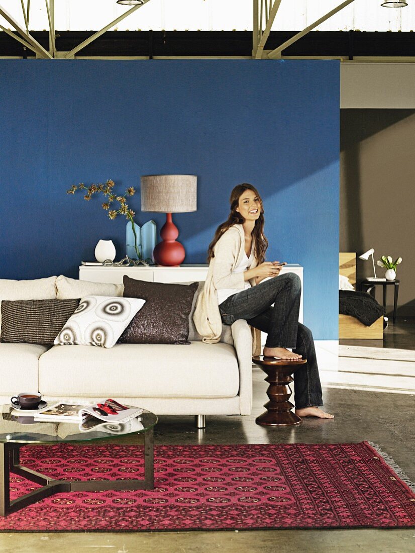 Wohnraum mit weißem Polstersofa, Orientteppich und Sideboard vor blauer Wand, Frau auf Armlehne des Sofas sitzend