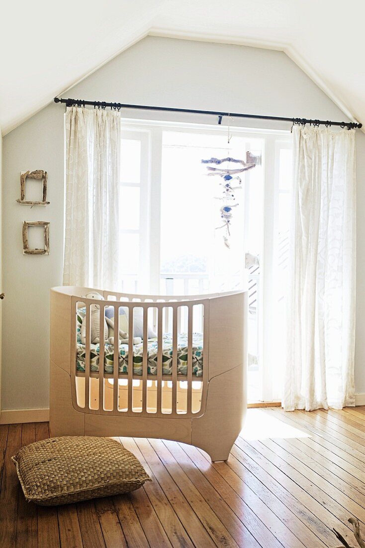 Custom-made, pale wood cot in front of balcony doors with airy curtains in attic nursery