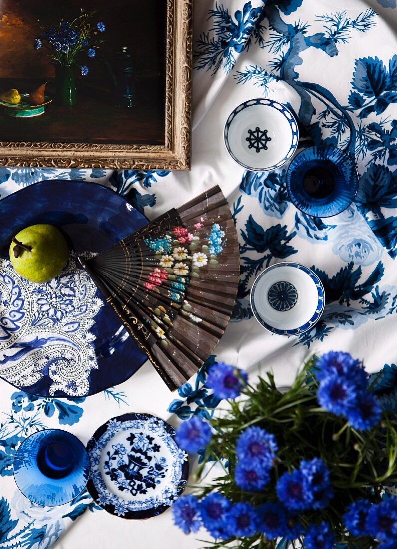 Blumenfächer und Schalen auf Tischdecke mit weiss-blauem Muster, vorne Kornblumen im Topf