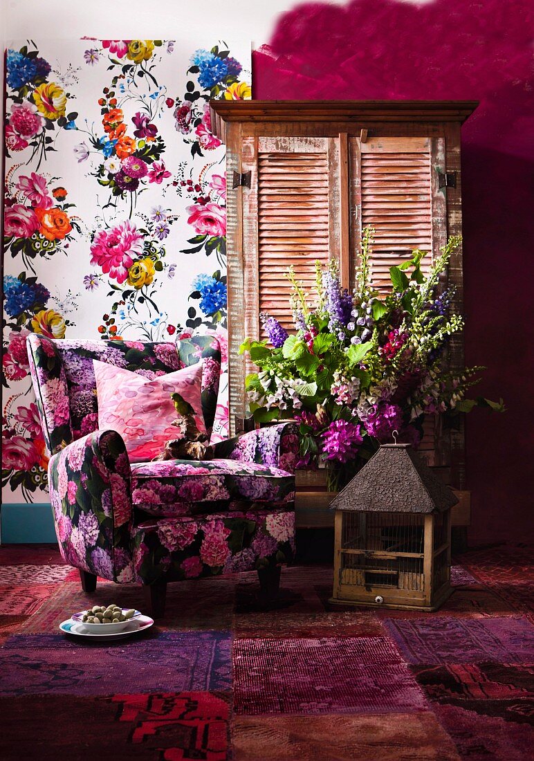 Floral sofa, old birdcage, patchwork rug in various shades of red, bouquet, cupboard with louver doors and floral wall hanging