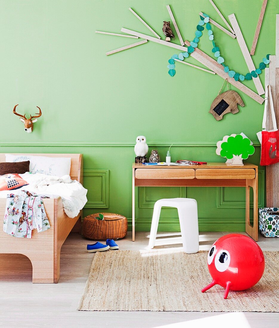 Modernes Holzbett neben Sekretär und weisser Kunststoff Hocker vor grüner Wand, daran montierter stilisierter Baum aus Holzleisten, vorne roter Hupfball auf Teppich