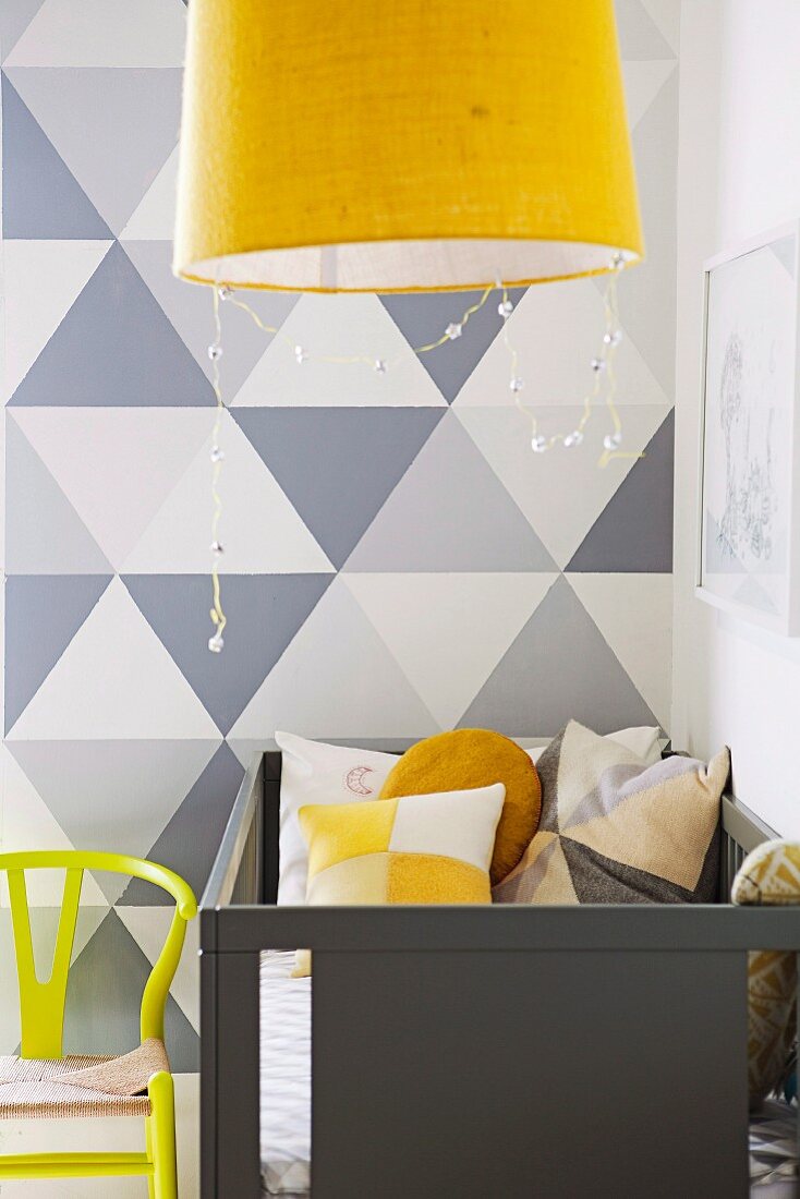 Pale grey cot against wall with painted grey geometric pattern and yellow pendant lamp in foreground