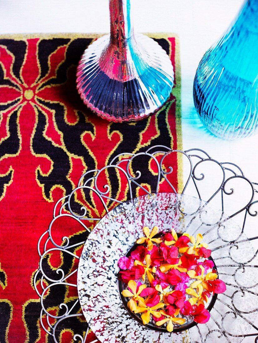 Artistic metal dish of red and yellow flowers and two shiny vases on rug with red and black pattern