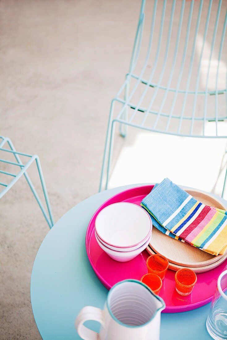 Pinkfarbenes Tablett mit Geschirr auf blauem runden Terrassentisch, im Hintergrund filigrane Metallgitter-Stühle