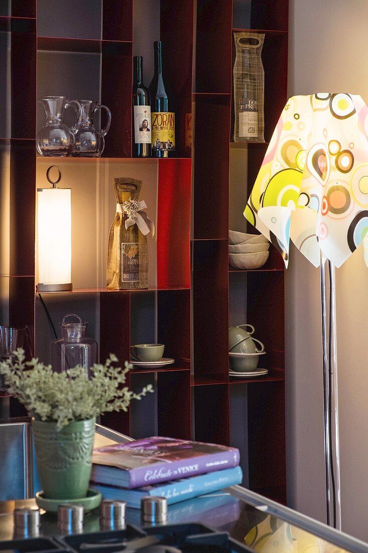 Standard lamp with retro lampshade in front of wine bottles and crockery on open-fronted shelves