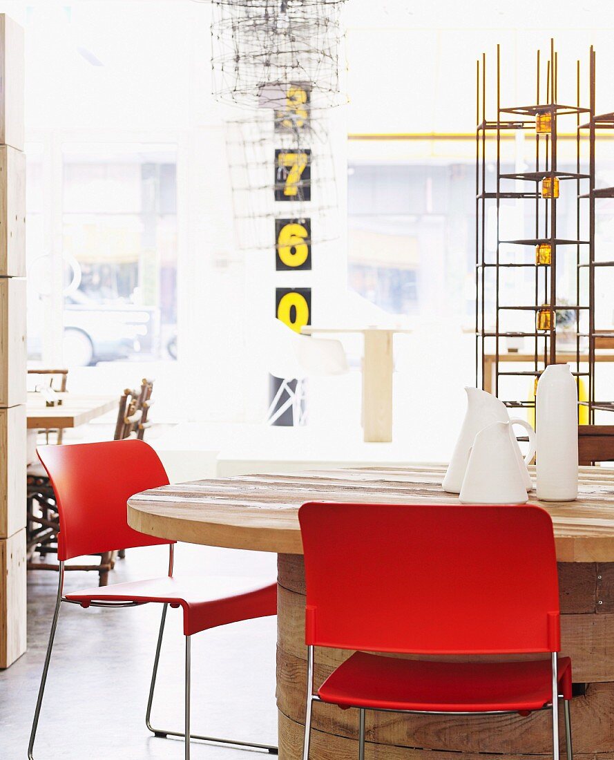Stühle mit roter Rückenlehne und Sitzfläche auf Metallgestell um runden Holztisch, darauf weiße Karaffen, im Hintergrund Regalgestell aus Metall