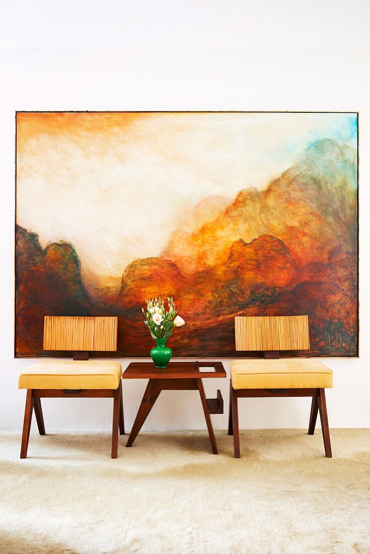 Designer wooden chairs with seat cushions an vase of flowers on side table in front of landscape painting