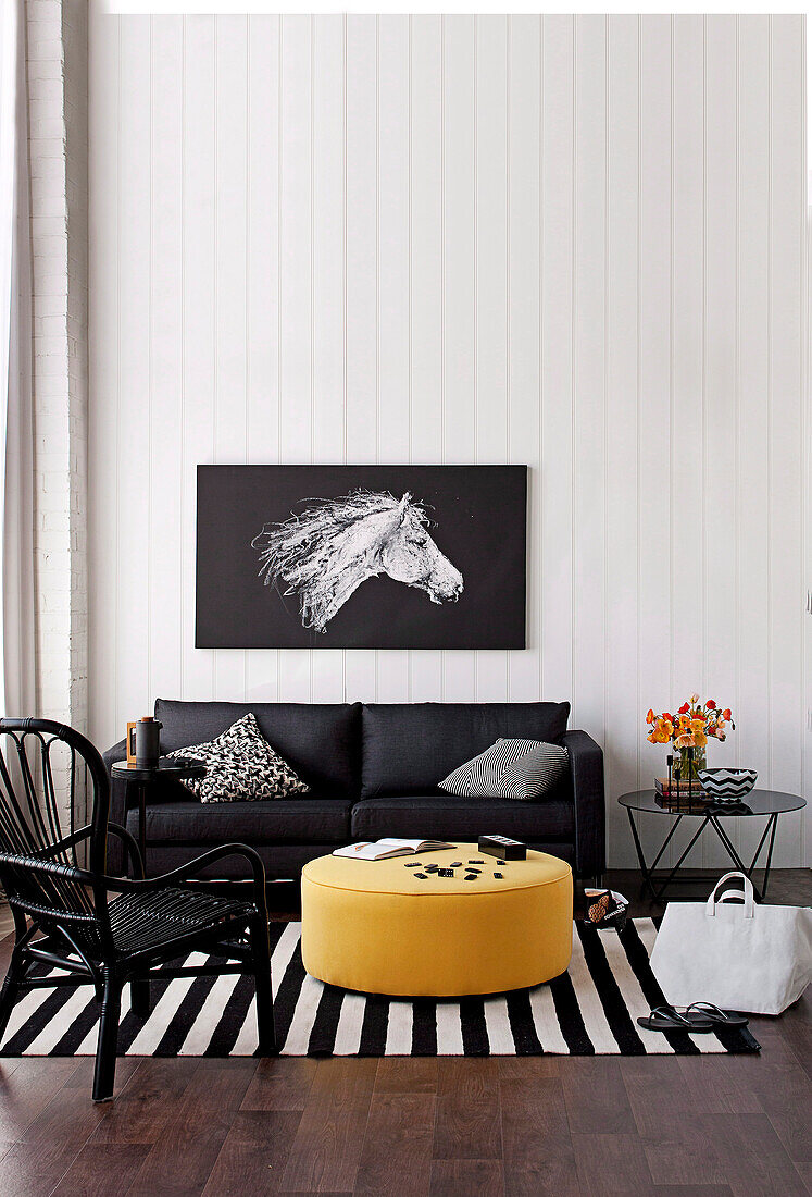 Rattan chair and black sofa around yellow upholstered coffee table on black and white striped carpet runner, on wall picture with horse's head