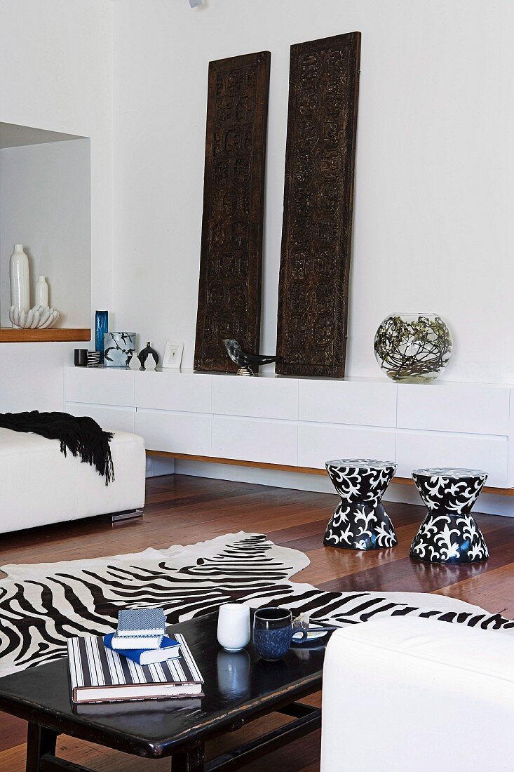 Weisser Lowboard mit Vasen und geschnitzten, indischen Holztäfeln, davor schwarz-weiße Hocker, Zebrafell und Couchtisch in hellem Wohnzimmer