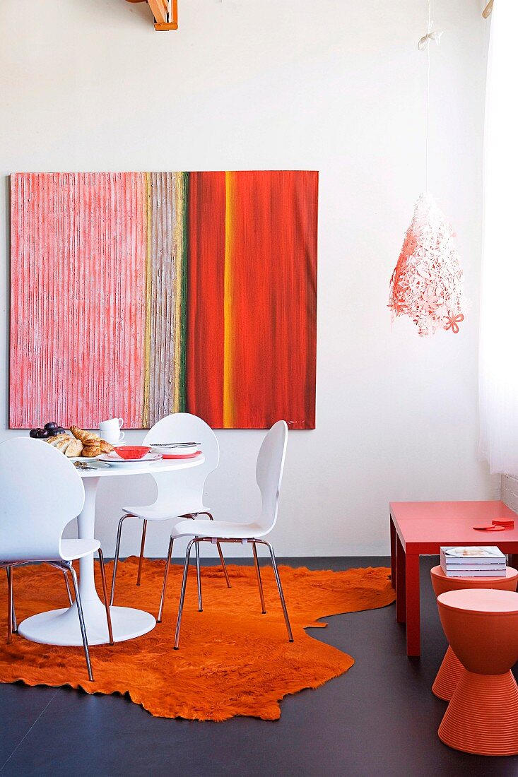 White shell chairs with metal frames and classic table on orange mock-animal-skin rug below modern picture on wall; orange stools and red side table to one side