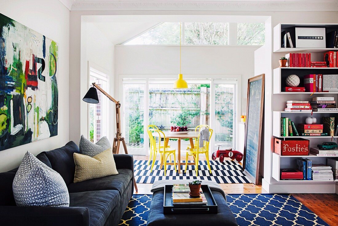 Open-plan interior - black couch, standard lamp with wooden base and coffee table on patterned rug; dining area with yellow-painted Thonet chairs in bright conservatory in background