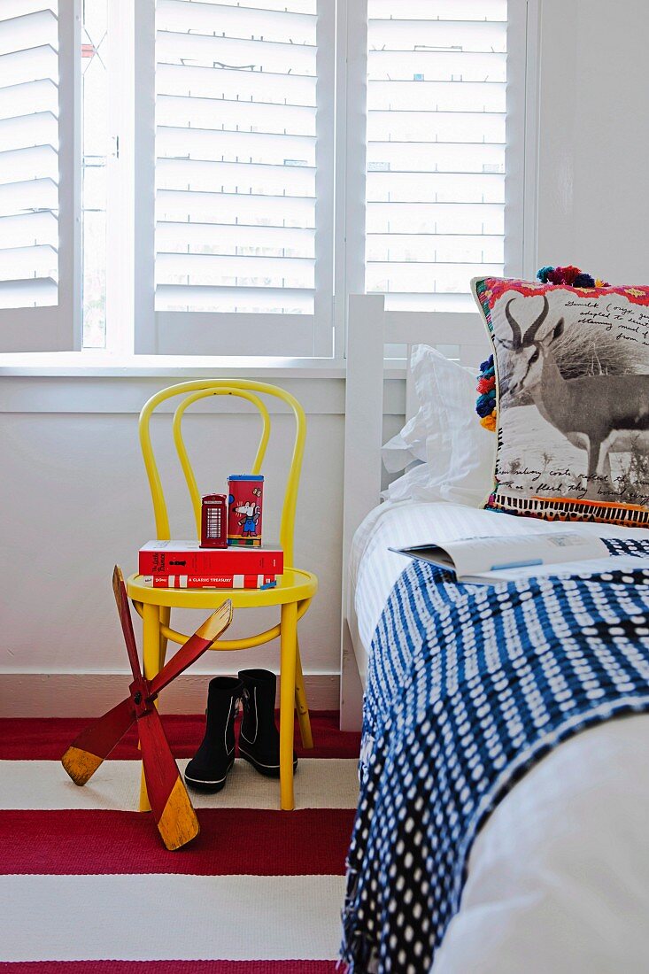 Detail of child's bedroom - toys on yellow-painted Thonet chair below window with closed white interior shutters and bed with patterned bedspread