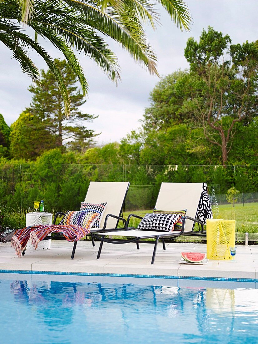 Two sun loungers with patterned cushions by the pool in front of glass balustrades and garden views