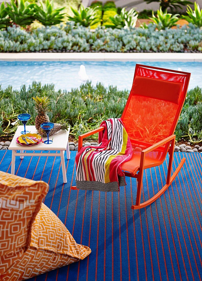 Orangefarbener Schaukelstuhl auf blauemTeppich neben Beistelltisch mit Cocktailgläsern vor Pool