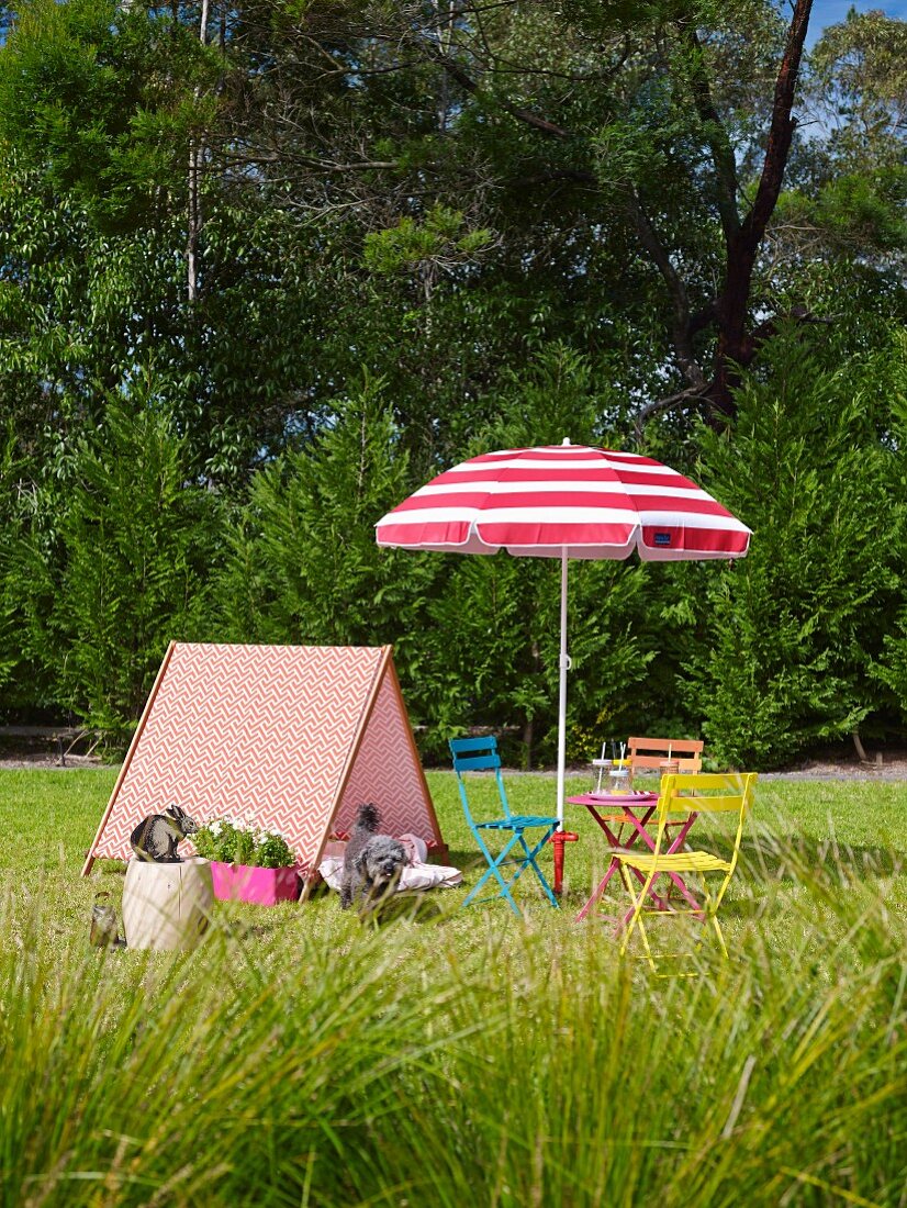 Zeltförmiges Holzgestell mit gemustertem Stoff bespannt neben bunten Gartenstühlen und rot-weißem Sonnenschirm