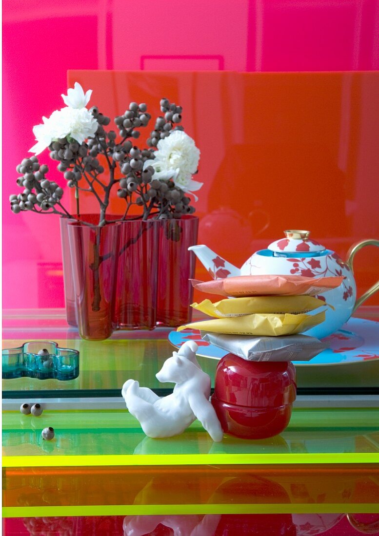 Buntes Stillleben mit roter Aalto Glasvase und edler Teekanne auf verschiedenen farbigen Glasplatten arrangiert