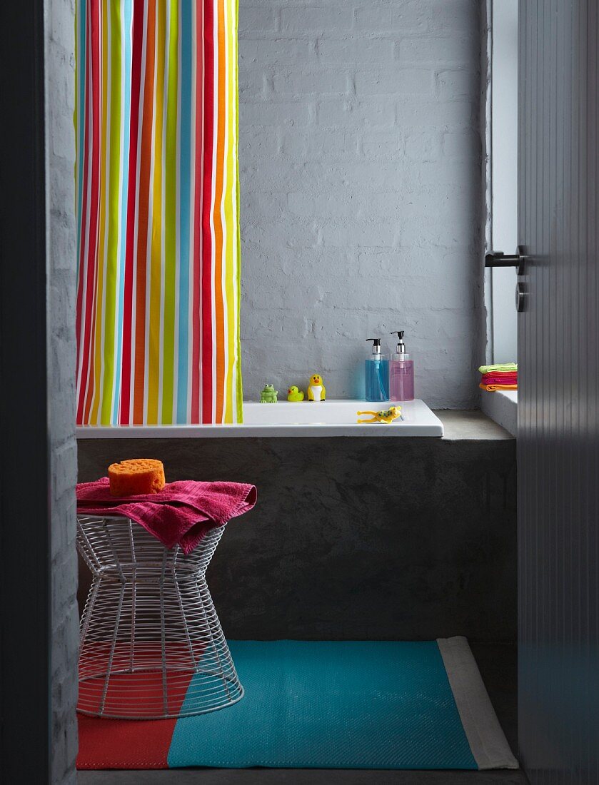 Blick auf bunt gestreiften Duschvorhang und Badewanne, Hocker aus Drahtgestell in grau getöntem Bad