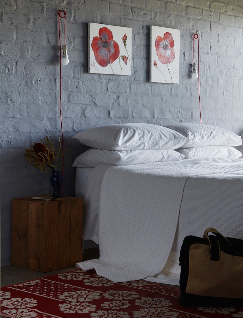 Doppelbett an grau gestrichener Ziegelwand mit rot-weißen Blumenbildern und innovativen Wandleuchten