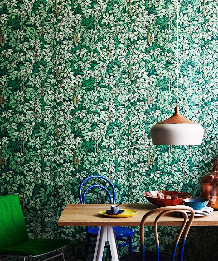 Retro lamp above dining table with Thonet chairs partially dipped in paint against leaf-patterned wallpaper