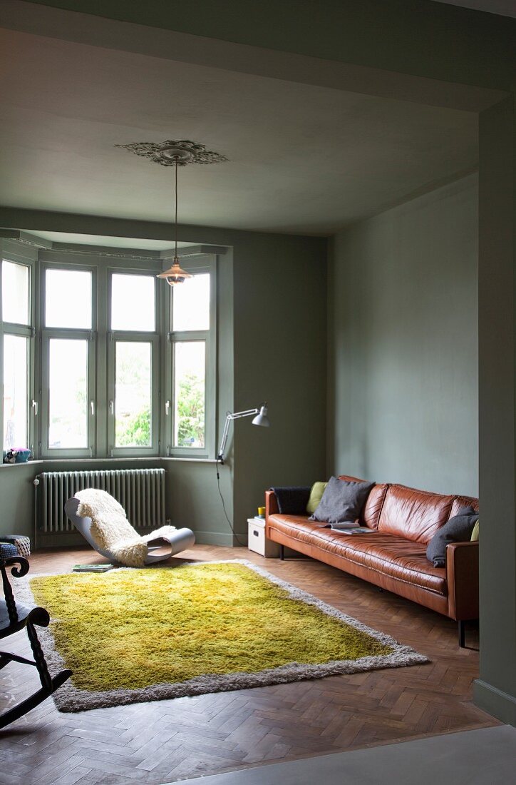 Erker-Wohnzimmer in dunklem Pastellgrün mit braunem Ledersofa, Designerschaukelstuhl und grünem Wollteppich in reduziertem Stil
