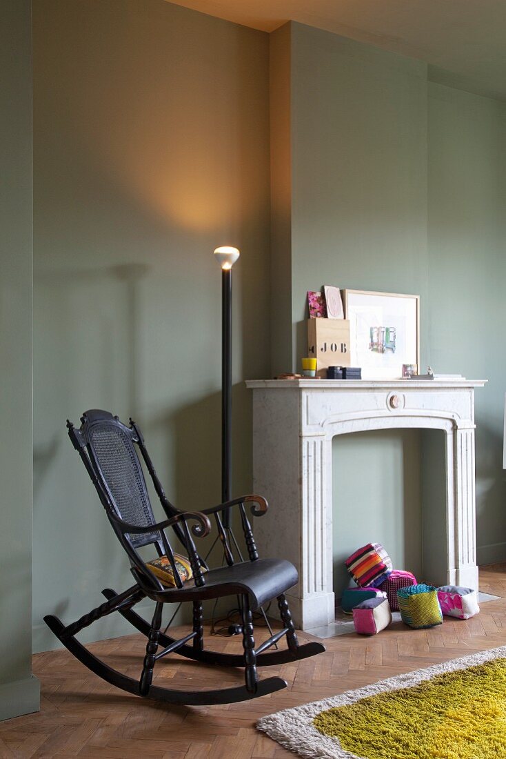 Nostalgic, black wooden rocking chair next to disused, decorated fireplace and illuminated standard lamp