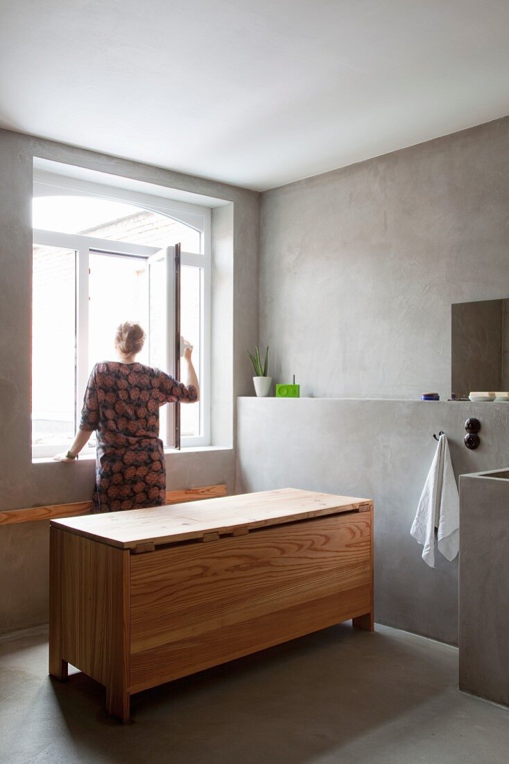 Designer wooden bathtub with closed lid on concrete floor in austere, minimalist bathroom; woman standing at window