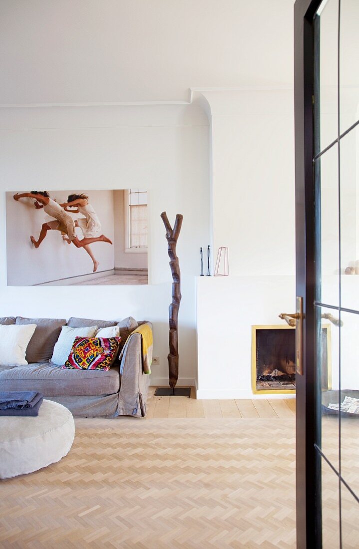 Blick durch offene Tür in minimalistischen Wohnzimmer mit Fischgrät-Parkettboden, neben offenem Kamin graue Polstercouch, an Wand grossformatige Photographie