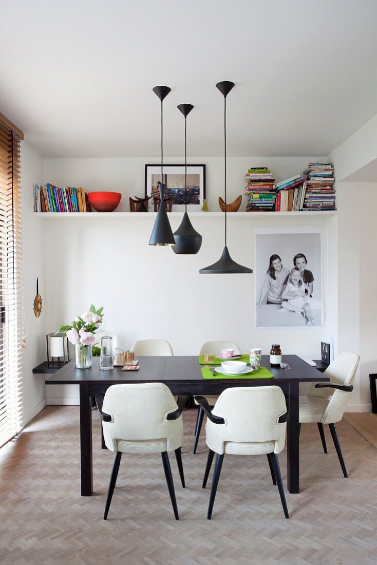Fifties-style chairs with white leather covers at black dining table below designer pendant lamps and family photo below shelves on wall in background
